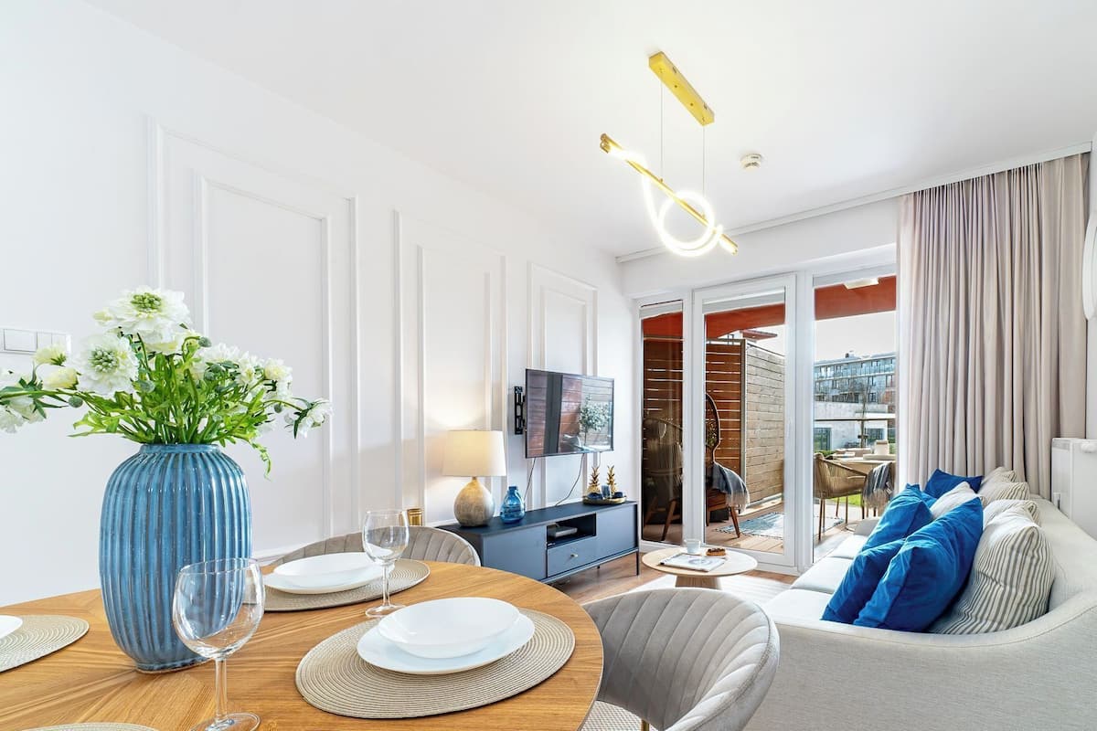 Ein modernes Wohnzimmer mit Essbereich, mit einer blauen Vase mit Blumen, einem runden Holztisch und einem grauen Sofa mit blauen Kissen.
