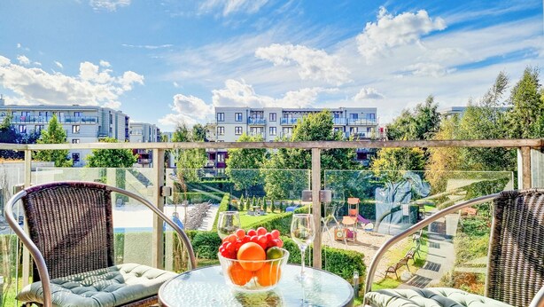 Un balcon avec des fauteuils en osier et une table sur laquelle sont disposés des fruits et un verre à vin, donnant sur un quartier résidentiel avec des immeubles modernes et de la verdure.