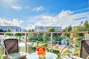 Een balkon met rieten stoelen en een tafel met fruit en een wijnglas, met uitzicht op een woonwijk met moderne gebouwen en groen.