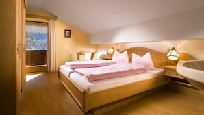 A bedroom with a wooden bed, two bedside tables with lamps, a window with curtains, and a view of snow-capped mountains. - Gasthof Dürrlehen (Berchtesgaden)