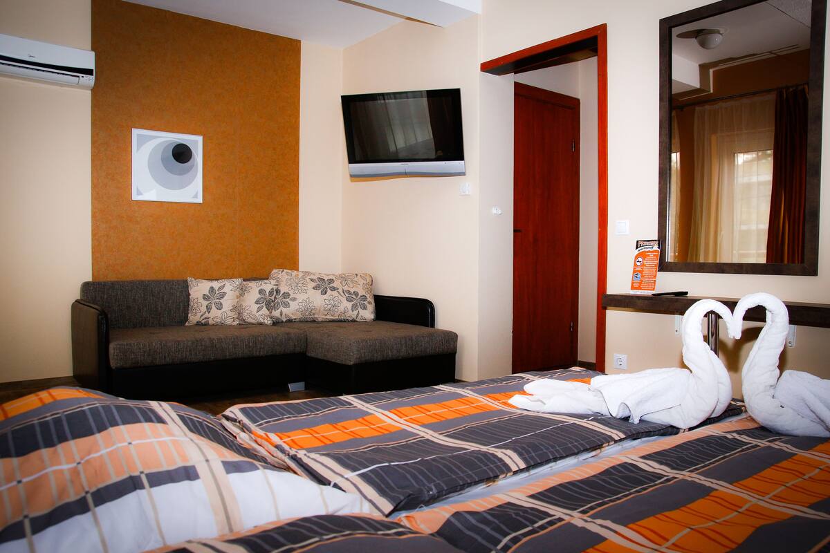 A hotel room with a sofa, a TV mounted on the wall, a mirror, and a bed with a decorative swan pillow arrangement.