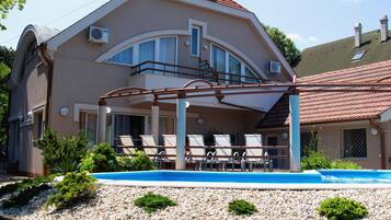 A two-story house with a balcony, a swimming pool, and a garden with pebbles and grass.