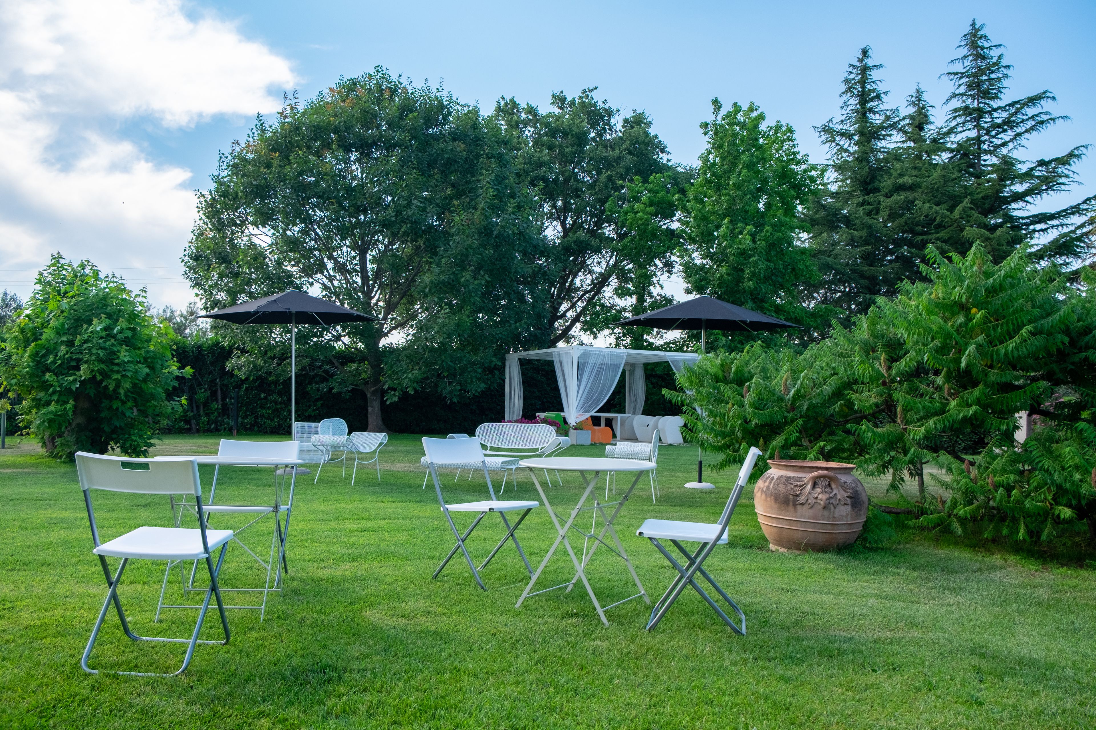 An outdoor seating area with white tables and chairs, a large potted plant, and a canopy structure.