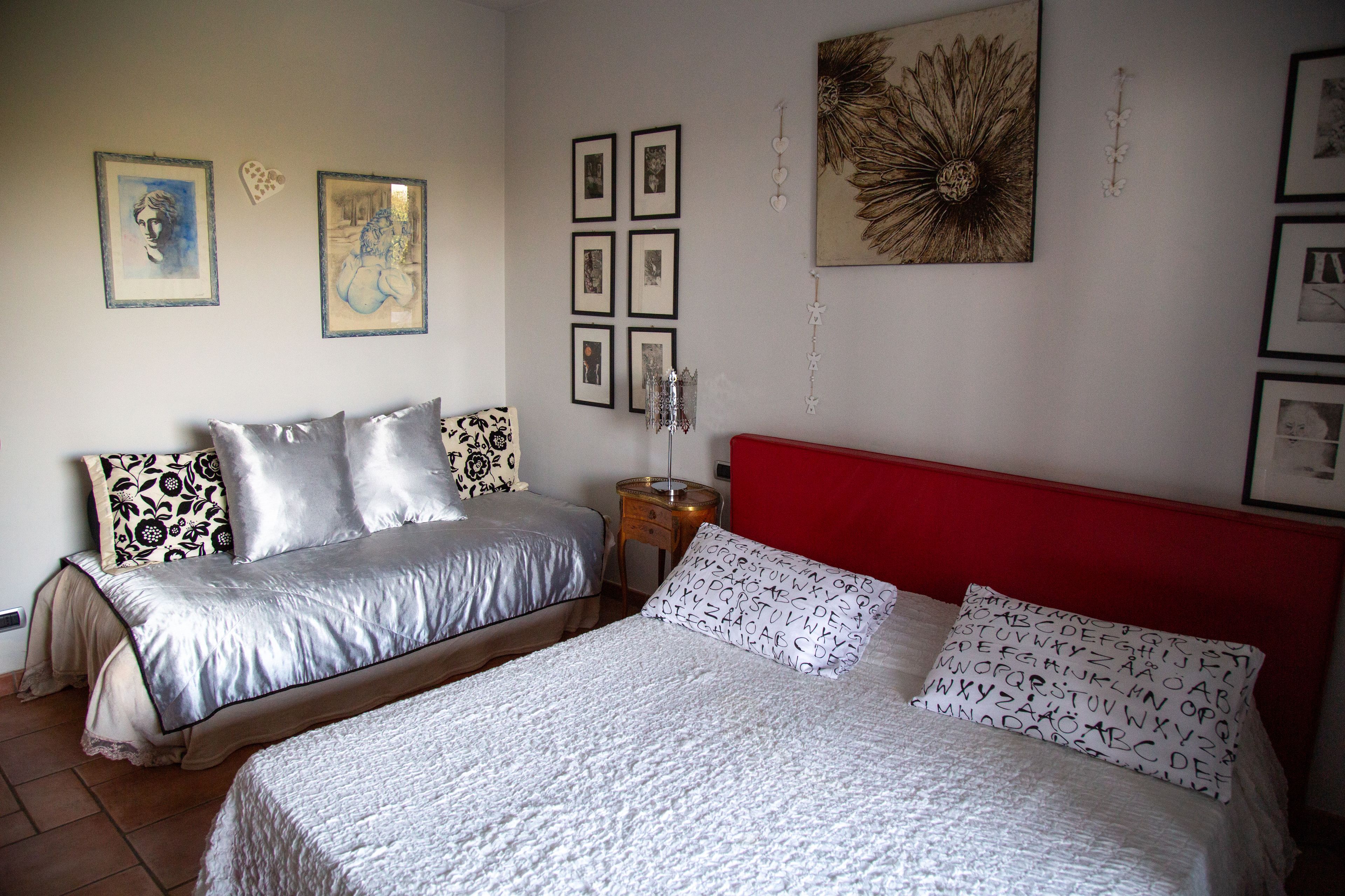 Ein Schlafzimmer mit einem roten Kopfteil, einem Doppelbett mit weißen und gemusterten Kissen, einem Einzelbett mit silbernem Kopfteil und einer Wand mit gerahmten Bildern und einem dekorativen Element.