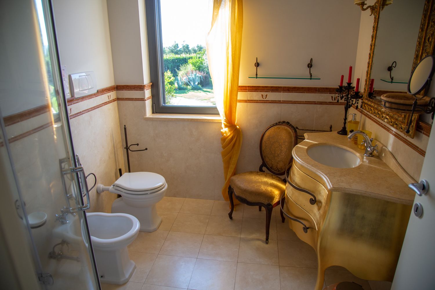 Une salle de bain comprenant des toilettes, un bidet et un lavabo. On y trouve également une chaise et une fenêtre avec des rideaux.
