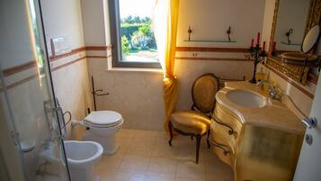 Une salle de bain comprenant des toilettes, un bidet et un lavabo. On y trouve également une chaise et une fenêtre avec des rideaux.