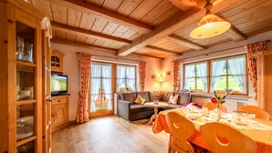 Een gezellige kamer met een houten plafond, een bank, een eettafel gedekt voor vier personen en een televisie op een kast.