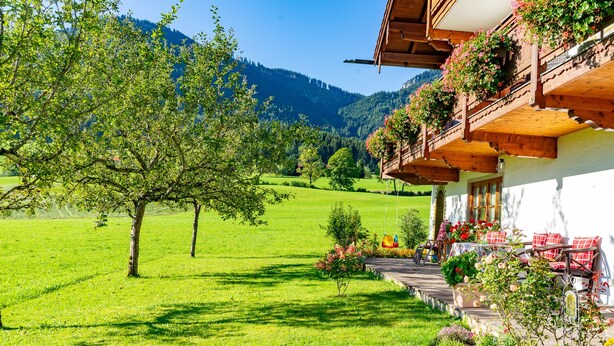 Een klassiek chalet met een balkon versierd met bloemen, een verzorgde tuin en uitzicht op een groen dal en bergen.