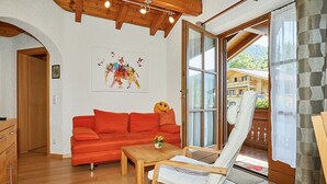 Een gezellige kamer met een houten plafond, een rode bank, een houten tafel en een witte stoel. Er is een groot raam met uitzicht op gebouwen en groen.