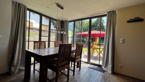 A dining area with a wooden table and chairs, large windows, and a view of an outdoor patio with yellow chairs and a red umbrella.