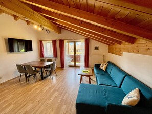A cozy room with a wooden ceiling, a dining table with chairs, a sofa, and a television mounted on the wall.