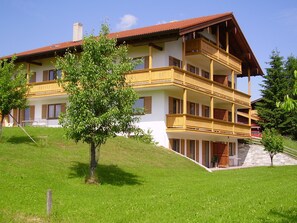 A multi-story building with balconies, a sloped roof, and a tree in the foreground.