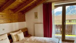 A bedroom with a wooden ceiling, a bed with white linens, a radiator, and a view of a mountain through a window with red curtains. - Vacation apartment A, 4 pers., panoramic balcony, bedroom, bed-sitting room, 67 sqm (Inzell)