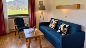 A living room with a blue sofa, a wooden coffee table, a floor lamp, and a view of a green field and mountains through a window. - Vacation apartment A, 4 pers., panoramic balcony, bedroom, bed-sitting room, 67 sqm (Inzell)