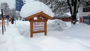Uma placa de madeira com o texto 'Haus am Maibaum Ferienwohnungen' parcialmente coberta por neve.
