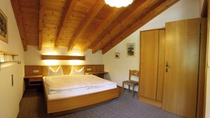 Quarto com teto de madeira, cama com roupa de cama branca, guarda-roupa de madeira e uma pequena cadeira.