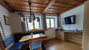 Ein gemütliches Zimmer mit Holzbalkendecke, einem Esstisch mit Stühlen, einer Bank, einem an der Wand befestigten Fernseher und einem Fenster mit Vorhängen.