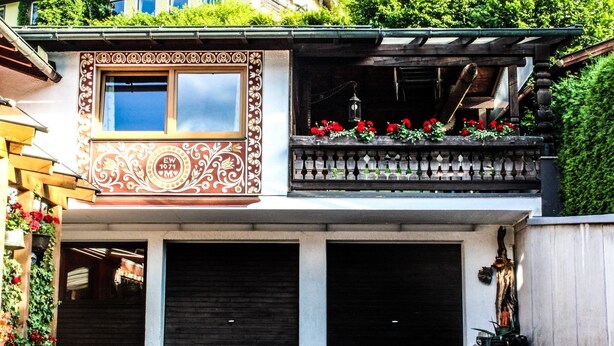 Ein zweistöckiges Gebäude mit einer verzierten Fassade, einem Balkon mit roten Blumen und zwei Garagentoren.