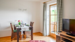 A dining area with a table set for two, a television on a stand, and a window with curtains. - Apartment Staufenblick for - 2 persons, 50 sqm, balcony with unobstructed view of the mountains (Bad Reichenhall)