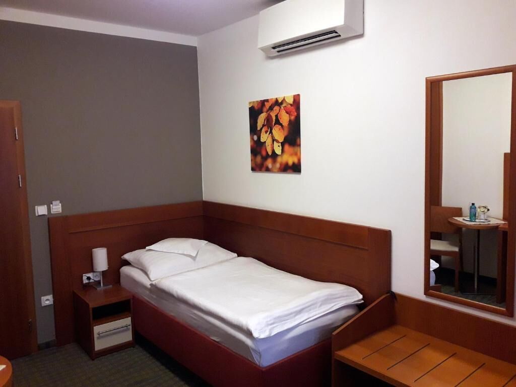 A neatly made bed with a wooden headboard, a bedside table with a lamp, a small drawer, and a wall-mounted air conditioner.