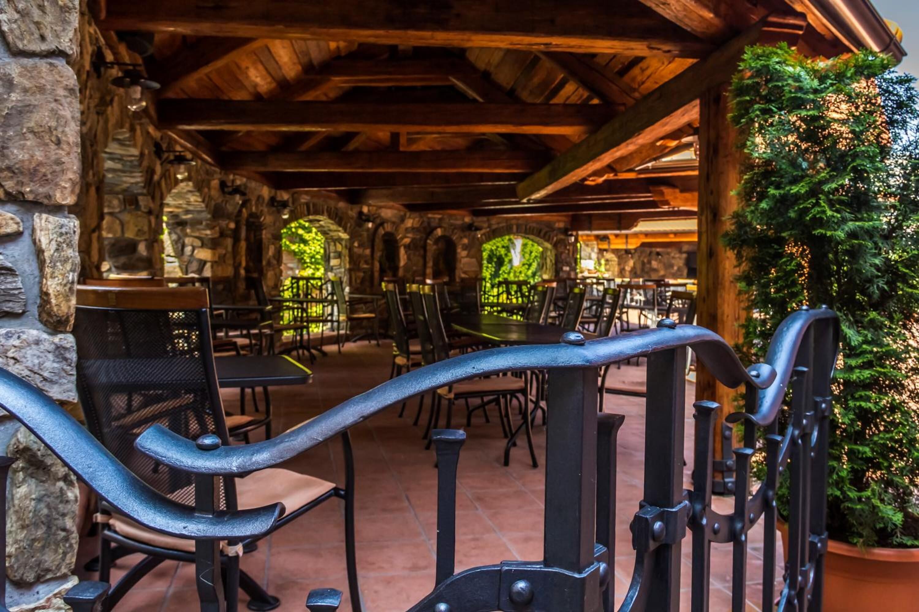 An outdoor patio area with stone walls, wooden beams, and metal railings. Tables and chairs are arranged for dining.