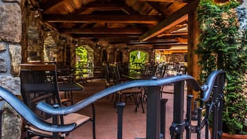 An outdoor patio area with stone walls, wooden beams, and metal railings. Tables and chairs are arranged for dining.