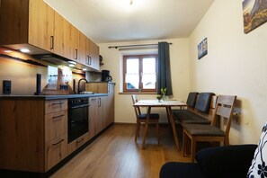 A compact kitchen with wooden cabinets, a dining table with chairs, and a window with curtains.