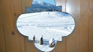 Eine Holzkonstruktion mit einem runden Fenster, das eine verschneite Landschaft mit Bäumen und einem Berg im Hintergrund zeigt.