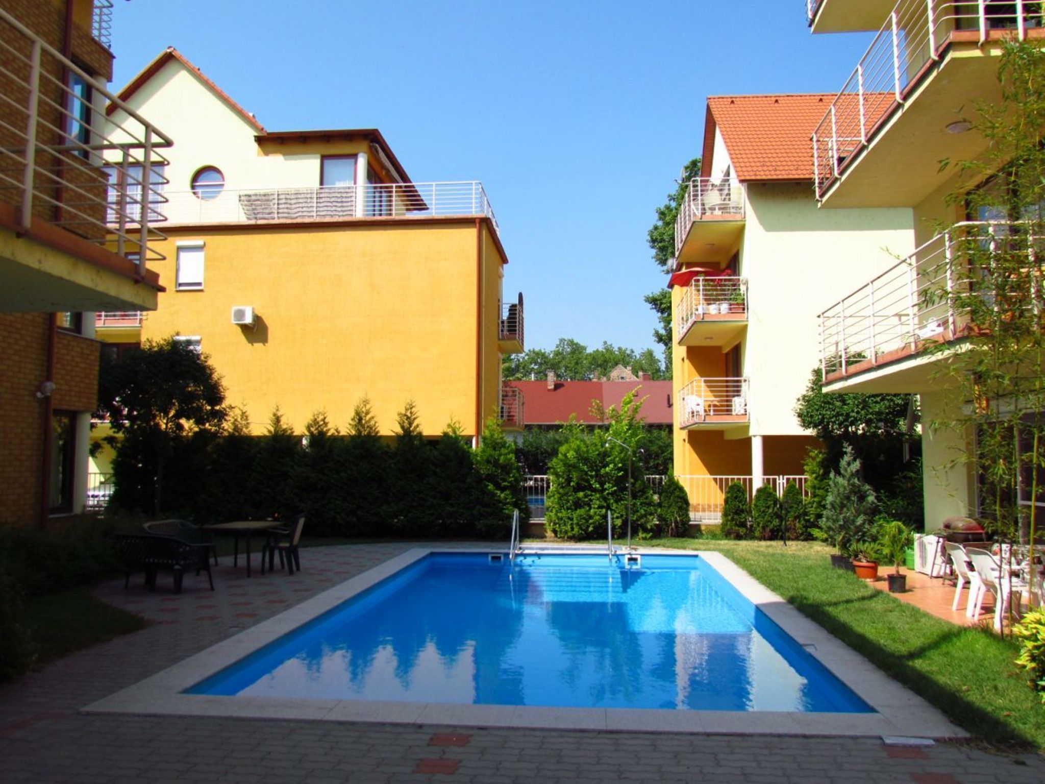 A rectangular swimming pool with a clear blue water surface, surrounded by a paved area with a few chairs and tables.