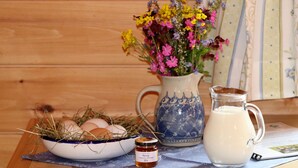 A wooden table with a bowl of eggs, a jar of honey, a pitcher of milk, and a vase of flowers.