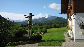 A rustic building with a paved pathway leading to a grassy area, surrounded by lush greenery and a mountain backdrop.
