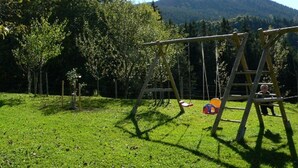Leikkikenttä, jossa on keinut ja liukumäki, sijaitsee nurmialueella, jossa on puita ja taustalla näkyy vuori.