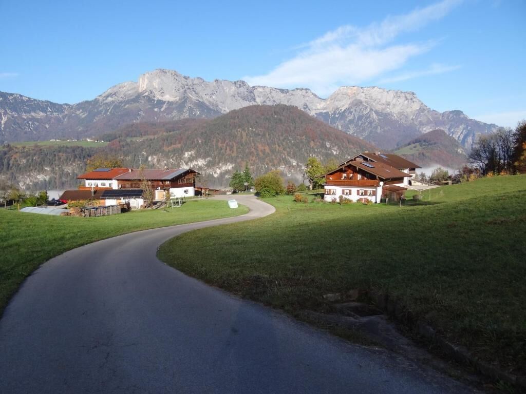 A rural road curving through a green landscape with traditional houses and a mountain backdrop.