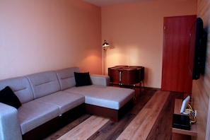 Uma sala de estar moderna com um sofá seccional cinza, piso de madeira, luminária de pé, mesa de madeira pequena e uma TV fixada na parede.