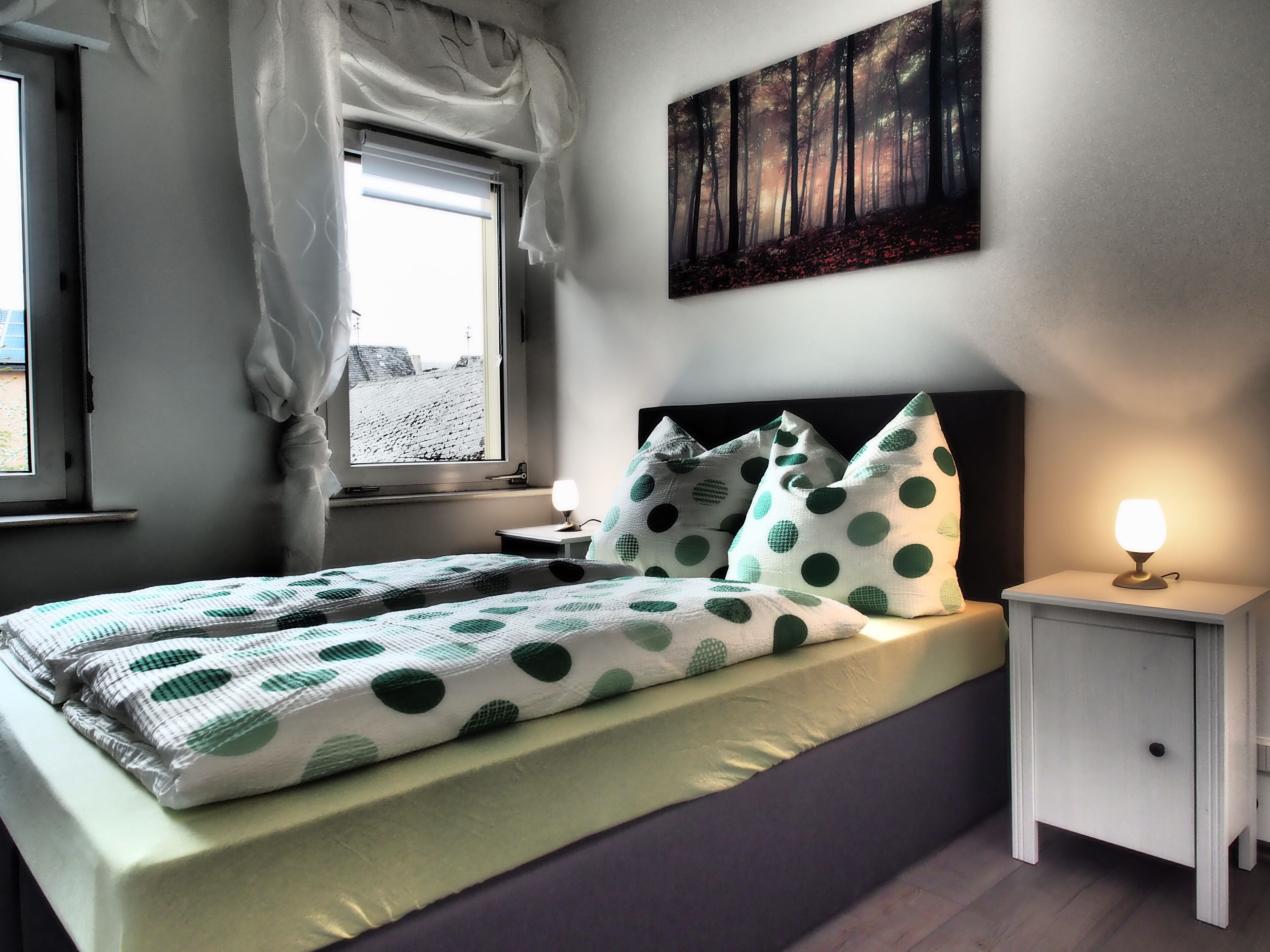 Et soveværelse med seng, puder, et sengebord med lampe, et vindue med gardiner og et billede på væggen.
