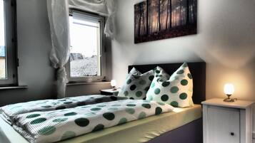 Et soveværelse med seng, puder, et sengebord med lampe, et vindue med gardiner og et billede på væggen.