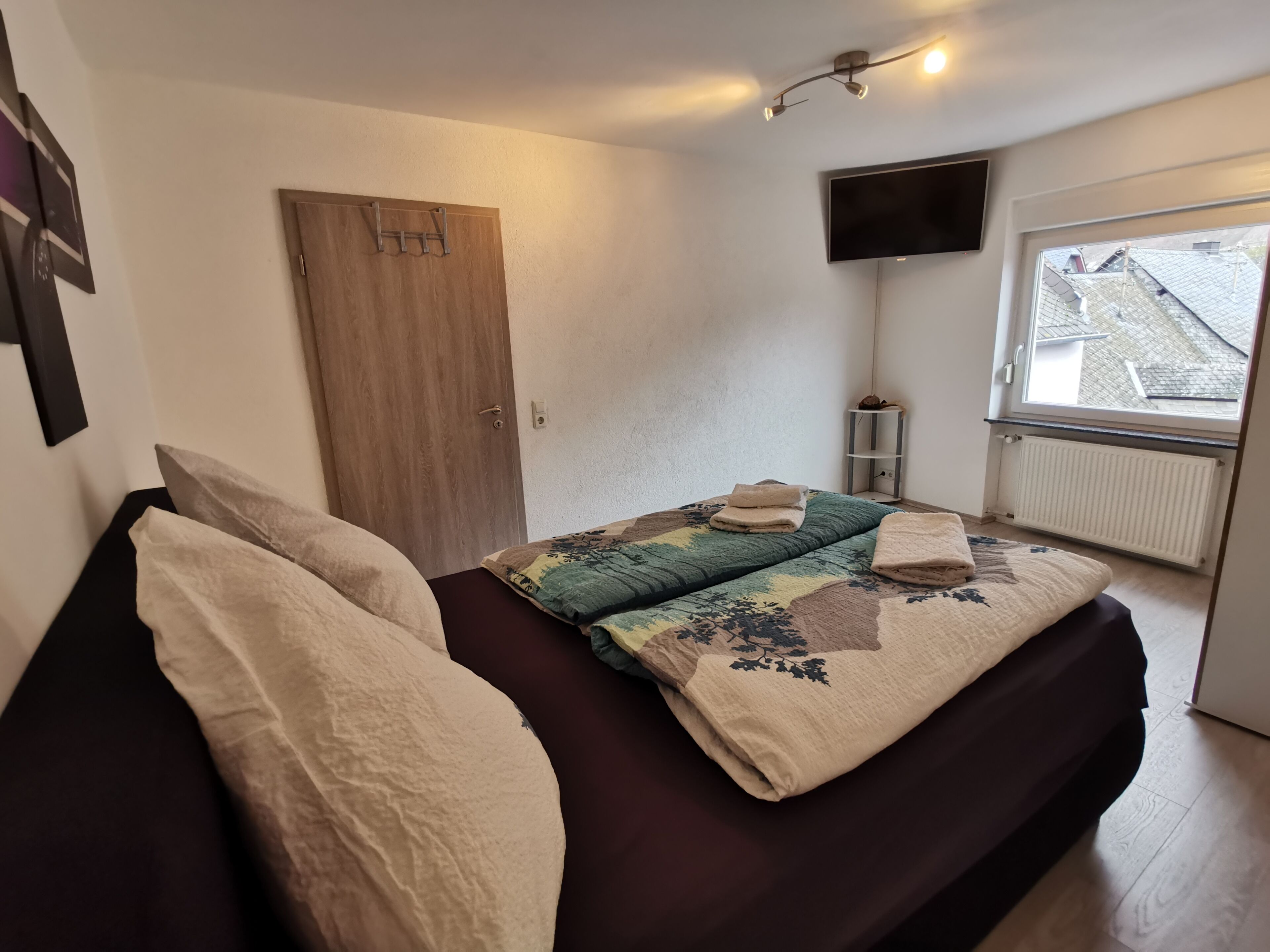 A bedroom with a bed, a wooden door, a TV mounted on the wall, and a window with a view of the outside.