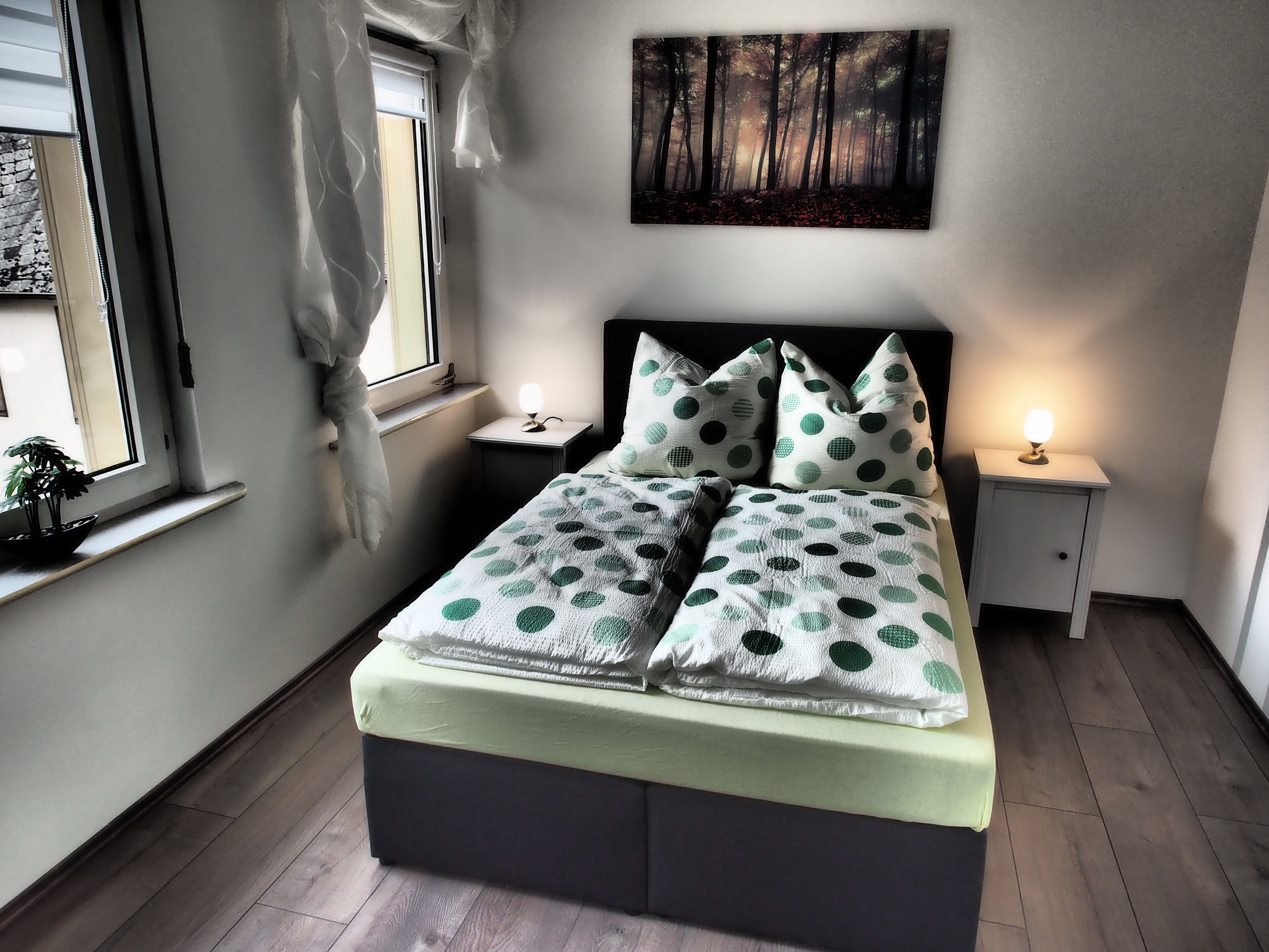 A bedroom with a bed, two bedside tables, a nightstand, a window with curtains, and a painting on the wall.