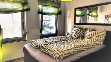 A bedroom with a bed, a window with green curtains, a bedside lamp, and a mirror on the wall.
