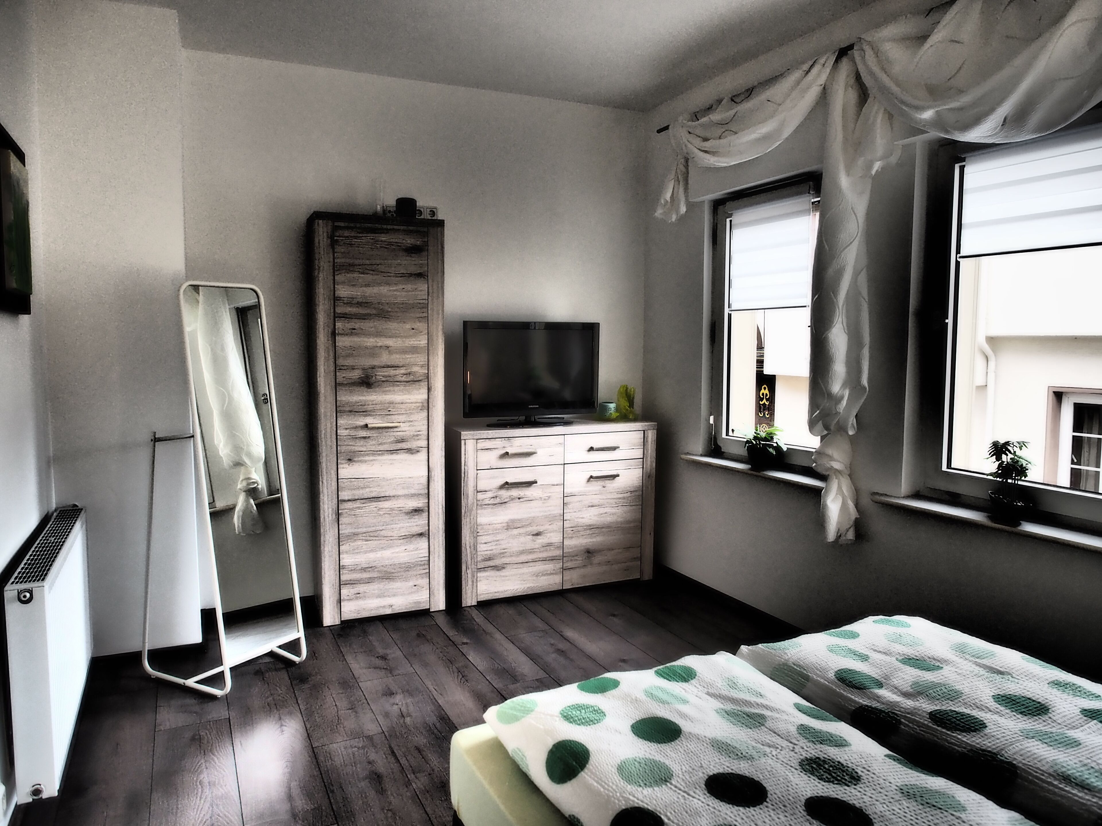 Une chambre avec une armoire en bois, un téléviseur à écran plat, un lit recouvert d’une couverture à pois, une fenêtre avec des rideaux et un radiateur.