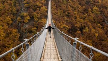 A suspension bridge with a person walking on it, surrounded by autumn foliage.