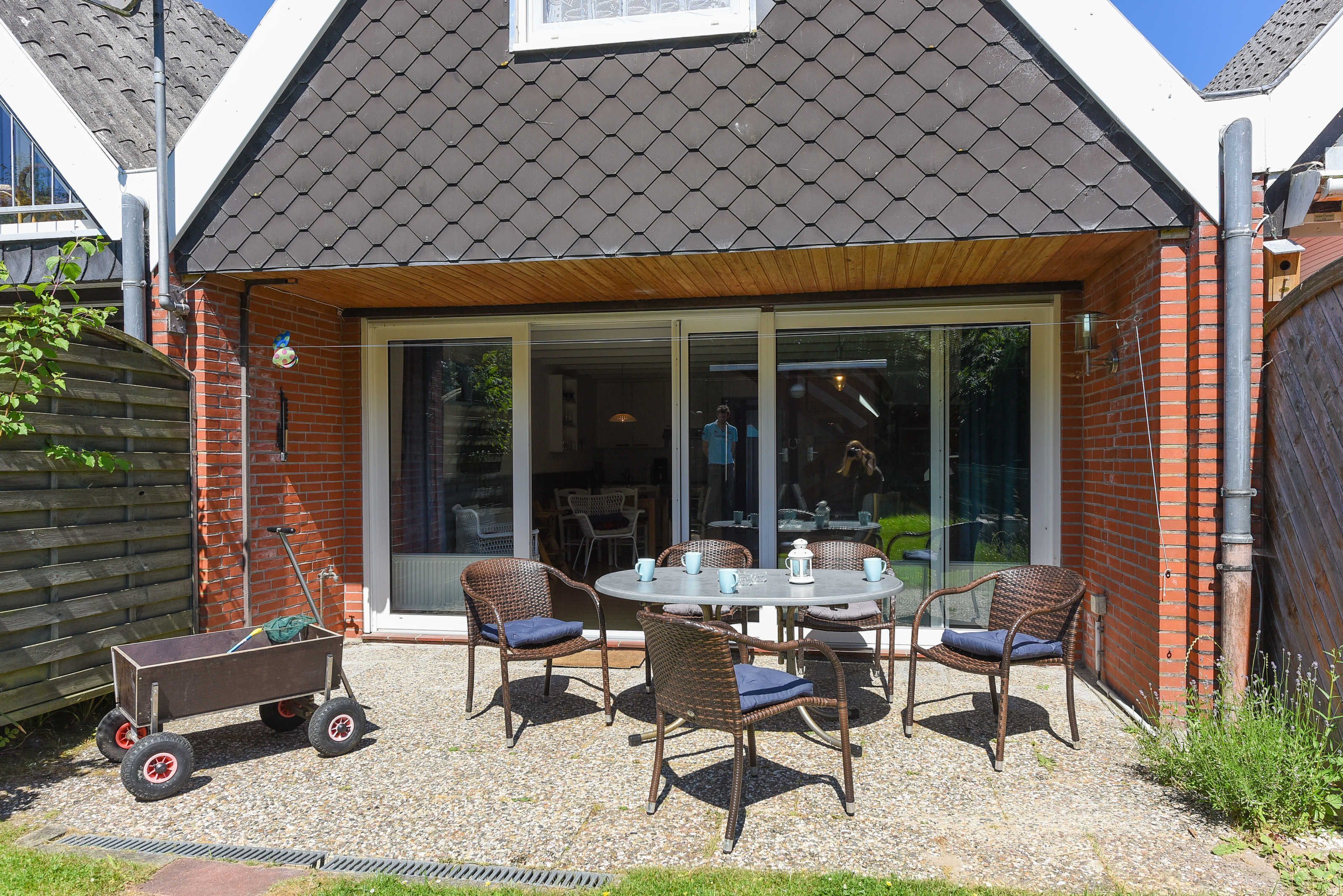 A patio with wicker chairs and a table, a wooden cart, and a house with a tiled roof and large glass doors.