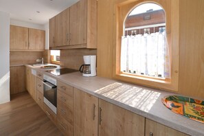 A kitchen with wooden cabinets, a built-in oven, a coffee maker, and a window with a curtain. - Vacation apartment Faerber 1, ground floor Dorfstr. 35 (Reit im Winkl)