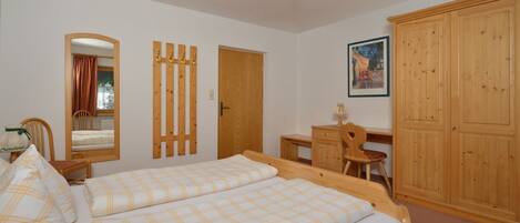 Een slaapkamer met een tweepersoonsbed, een houten garderobe, een bureau met stoel, een spiegel en een lijst met een afbeelding aan de muur.
