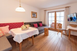 Un salon comprenant un canapé, une table agrémentée d’un centre de table floral, un tabouret en bois et un téléviseur diffusant une émission.