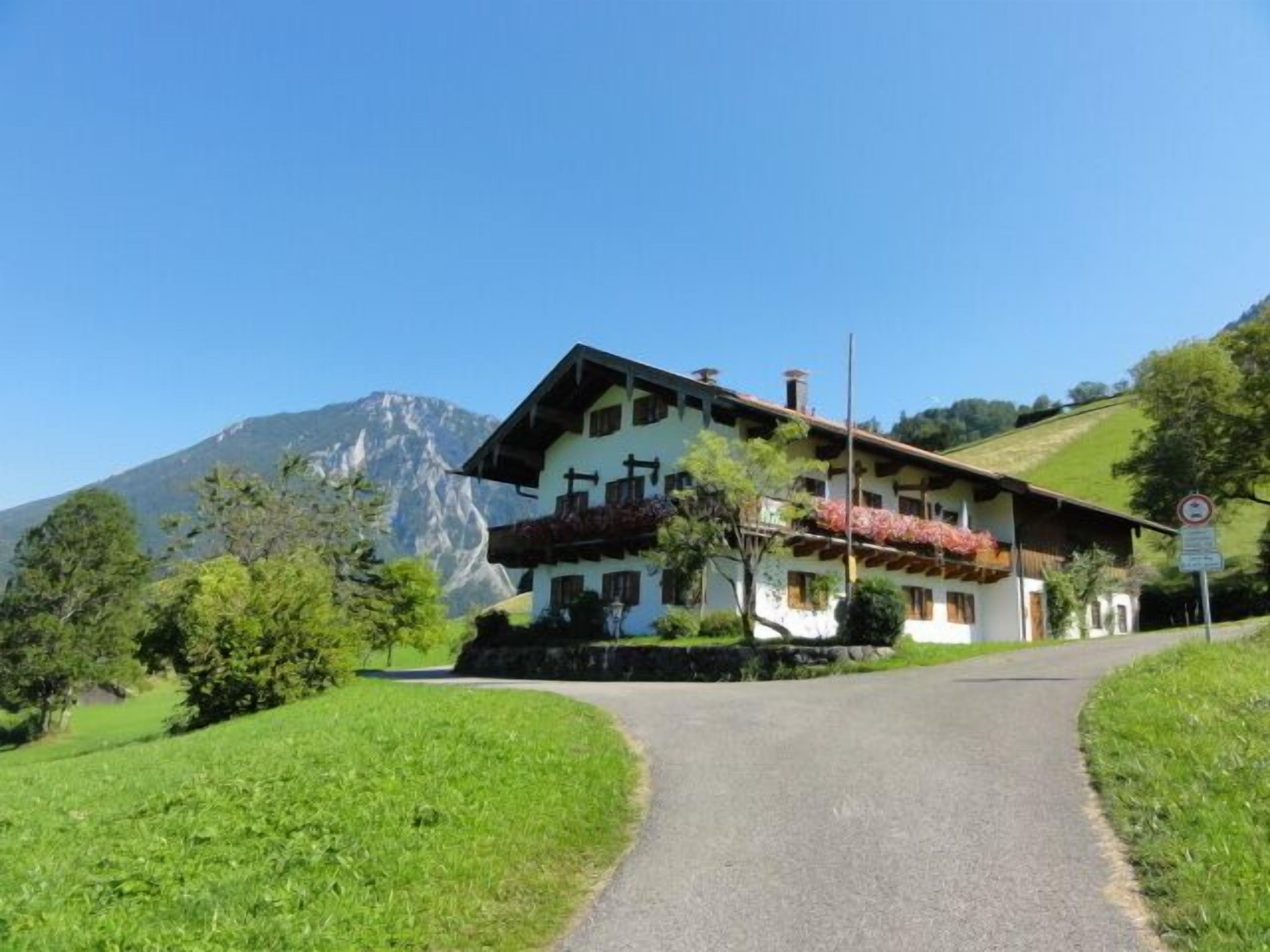 A traditional chalet with a sloped roof, a balcony with red flowers, and a mountain backdrop.