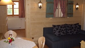 Una habitación acogedora con detalles de madera, un sofá azul, una mesa de comedor con un jarrón de flores y dos ventanas con cortinas a cuadros.
