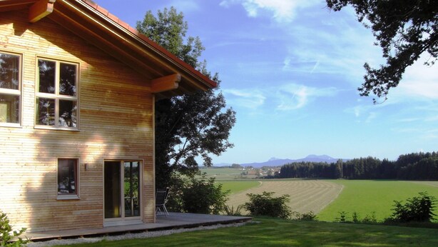 A wooden house with large windows and a view of a green field and mountains. - House Efeu 90m², up to 6 persons, 3 separate bedrooms (Traunwalchen)