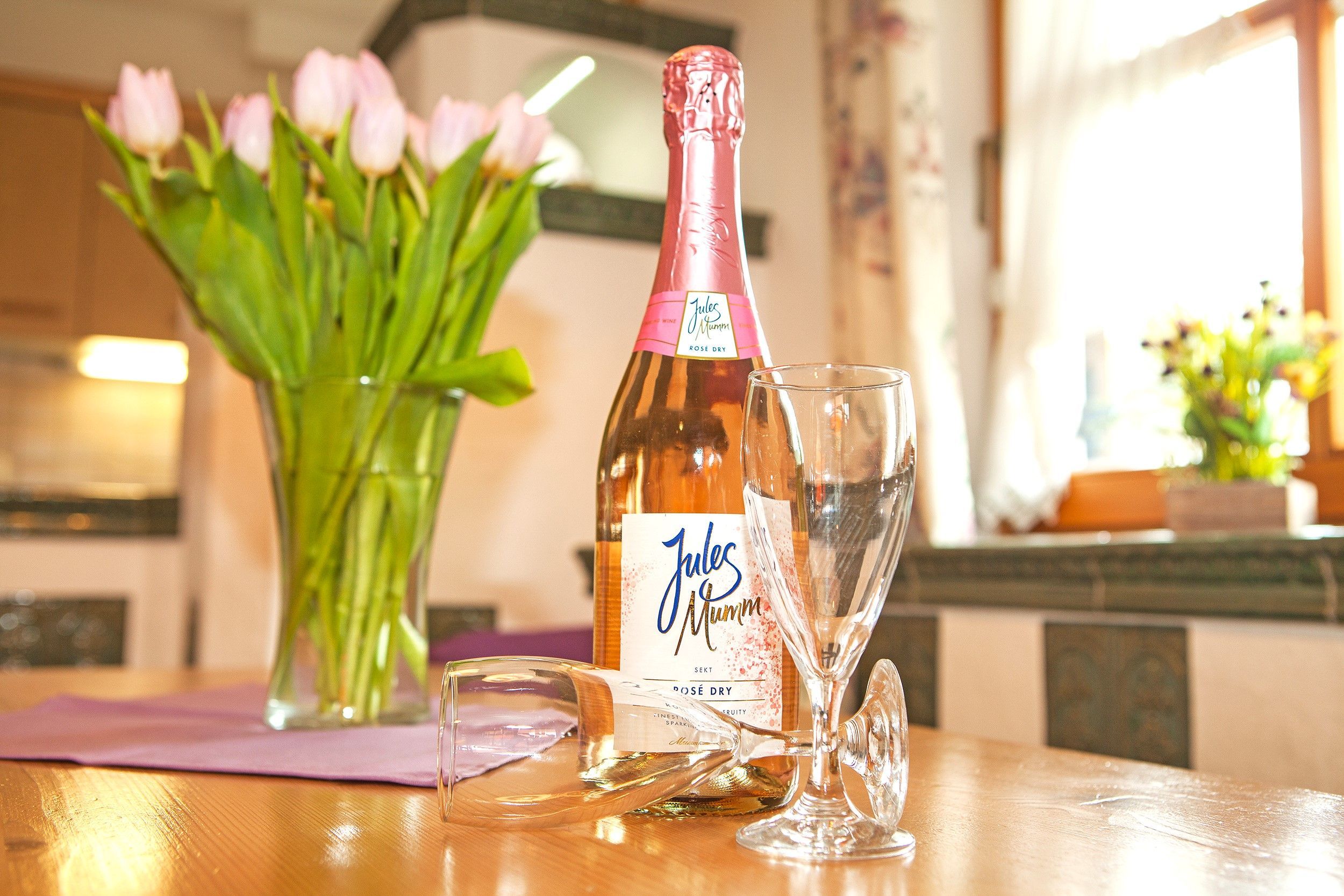 A bottle of Jules Mumm wine and a glass on a wooden table with a vase of flowers in the background.