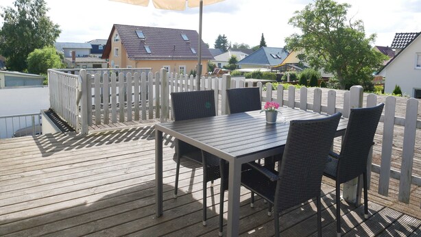 En udendørs terrasse med et bord og stole, en stor parasol og et hvidt stakittence.